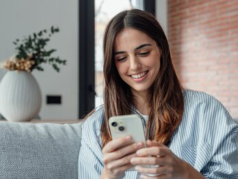 Smiling,Woman,Looking,At,Smartphone,Screen,,Sitting,On,Couch,,Enjoying